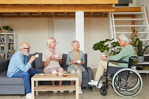 Group of senior friends talking to disabled man and drinking tea together in the living room