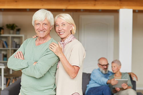 Portrait of happy senior couple smiling at camera while standing in the room with other couple in the background