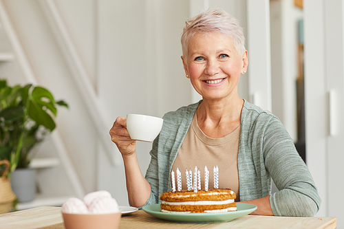 Happy senior woman smiling at camera while sitting at the table and drinking tea with cake