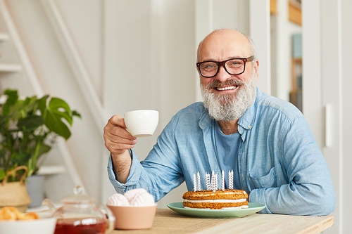 Portrait of senior bearded man in eyeglasses sitting at the table with birthday cake drinking tea and smiling at camera