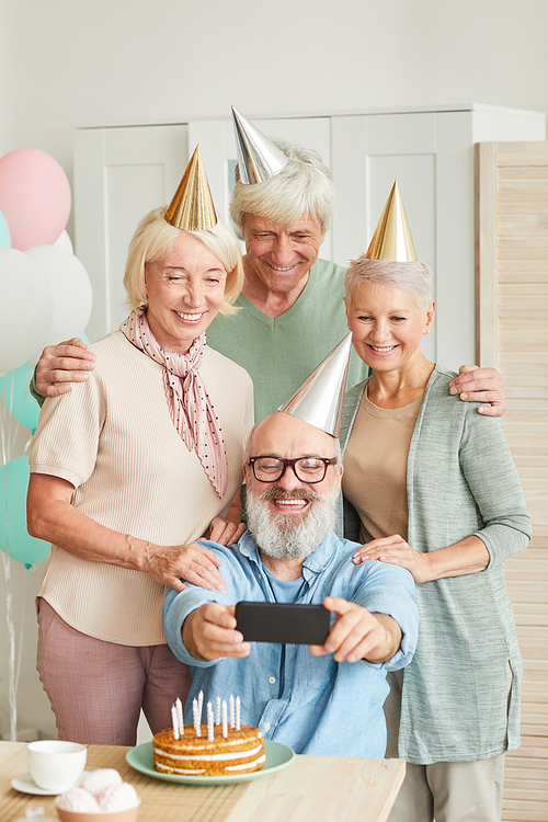 Group of senior friends in hats smiling at camera while making selfie on mobile phone during birthday party