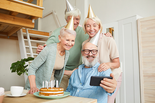 Senior people making selfie portrait on mobile phone while sitting at the table with birthday cake at party