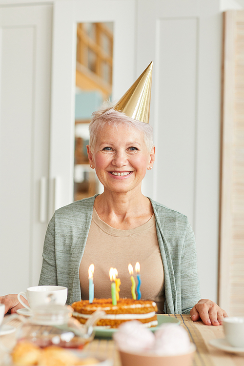 Portrait of happy senior woman in hat sitting at the table with birthday cake and smiling at camera at home