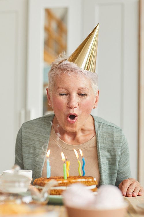 Senior woman in hat sitting at the table and blowing the candles on her birthday cake