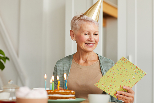 Senior woman sitting at the table with birthday cake reading a greeting card and smiling