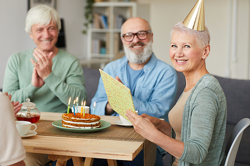 Portrait of happy senior woman reading greeting card and smiling at camera while celebrating birthday with her friends