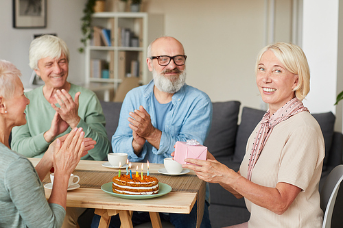 Portrait of senior woman holding gift box and smiling at camera with her friends sitting at the table with cake and applauding