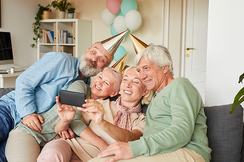 Senior people in party hats sitting on sofa and posing at camera together they making selfie portrait on mobile phone