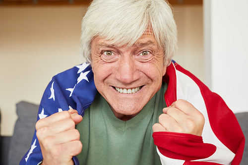Portrait of excited senior man with white hair with American flag on his back watching football at home