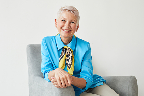 Portrait of elegant mature woman in beautiful blue blouse sitting on armchair and smiling at camera isolated on white background