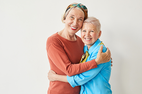 Portrait of two elegant mature women embracing each other and smiling at camera against the white background