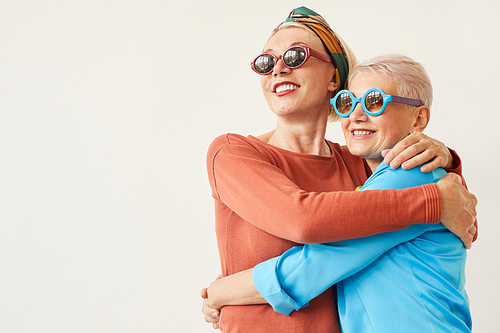Two happy senior women in sunglasses embracing each other while standing against the white background