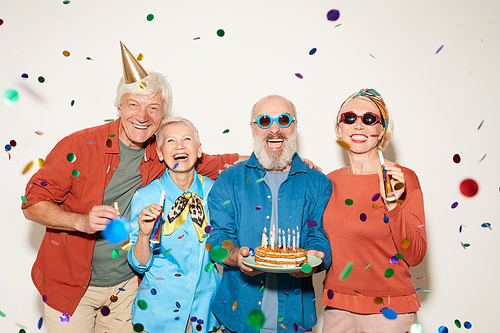 Portrait of group of senior people with birthday cake standing under the confetti and smiling at camera against the white background