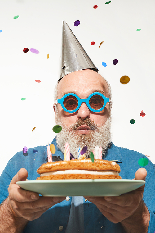 Portrait of senior man blowing the candles on his birthday cake under the confetti isolated on white background