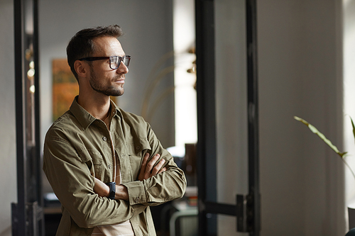 Young man in eyeglasses standing at office and looking through the window with pensive look