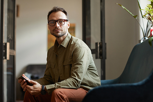 Portrait of young businessman in eyeglasses sitting on armchair with mobile phone and looking at camera at office