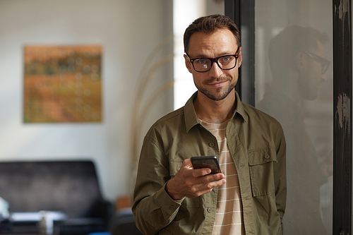 Portrait of young businessman in eyeglasses smiling at camera and typing a message on mobile phone at office