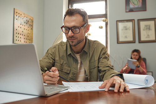 Mature businessman in eyeglasses sitting at his workplace in front of laptop and working over blueprint with his colleague in the background