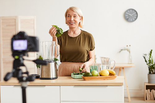 Smiling mature woman making a fruit cocktail and shooting this process on the camera in domestic kitchen