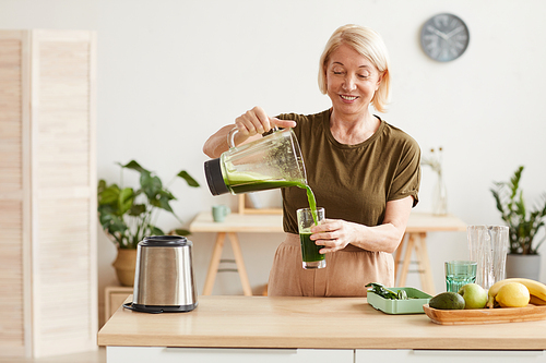 Smiling mature woman pouring fresh vegetable cocktail into the glass and drinking it