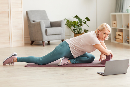 Senior woman doing exercises on exercise mat and watching these exercises on laptop at home