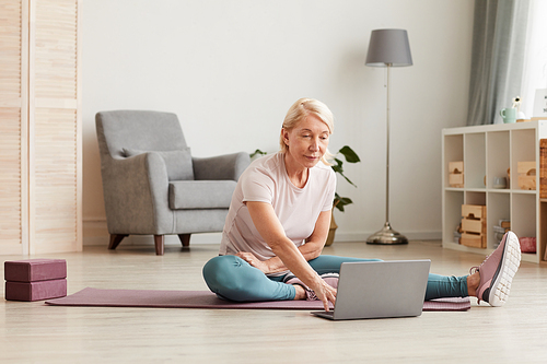 Mature woman sitting on the floor and using laptop computer during sports training at home