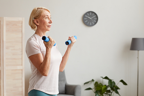 Senior woman with blond short hair holding dumbbells in her hands and exercising while standing in the room