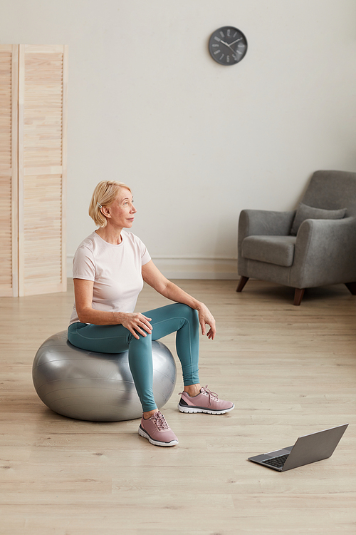 Mature woman sitting on the ball in front of laptop computer and exercising in the room