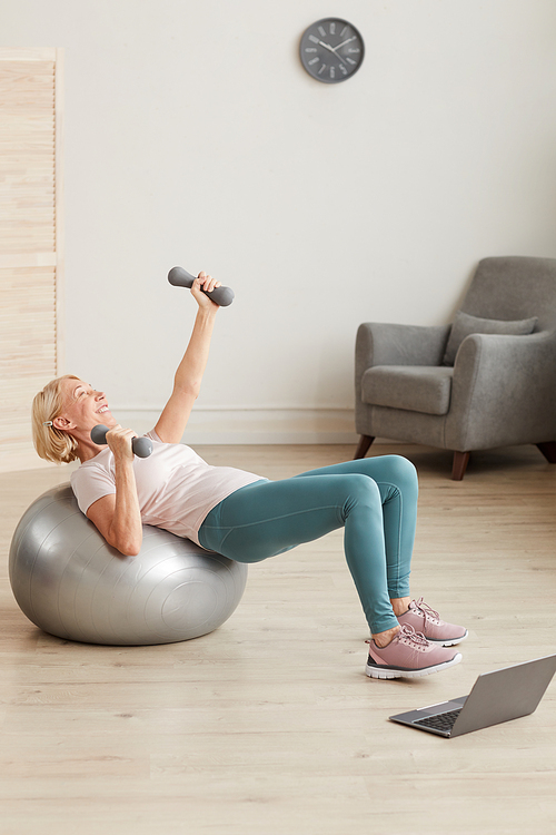 Mature woman exercising on the ball with dumbbells during online training at home