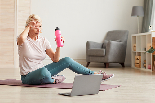 Mature woman sitting on the floor on exercise mat and drinking water after sports training at home