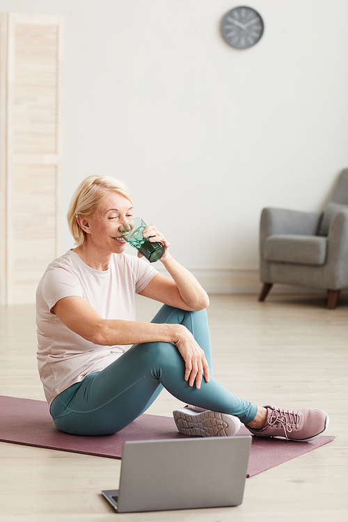 Senior woman sitting on the floor on exercise mat and drinking water after online training at home