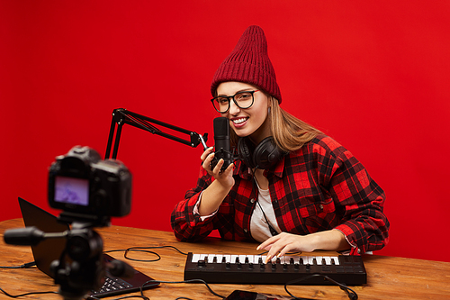 Young blogger sitting at the table playing on musical keyboard and singing in microphone she shooting video for her followers