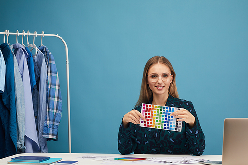 Portrait of young woman sitting at the table showing colored pattern and smiling at camera while working at studio