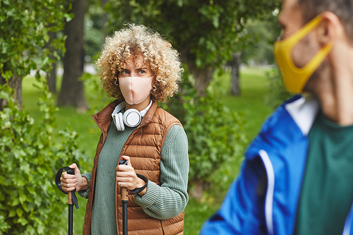 Young woman with curly hair in protective mask using sticks during sport walk with young man outdoors