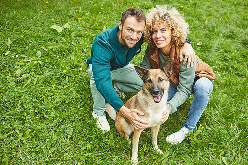 Portrait of young happy couple sitting on green grass with German shepherd and smiling at camera
