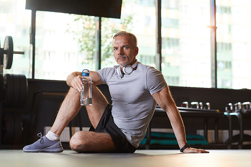 Portrait of mature man sitting on the floor and drinking water from the bottle he resting after sports training in gym