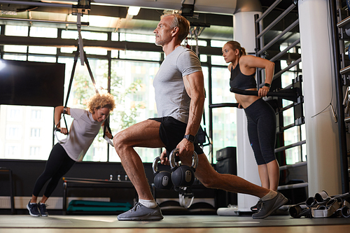 Mature man exercising with dumbbells in ym with two women in the background