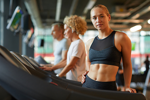 Portrait of beautiful sportive woman looking at camera during her training on treadmill with other people in the background