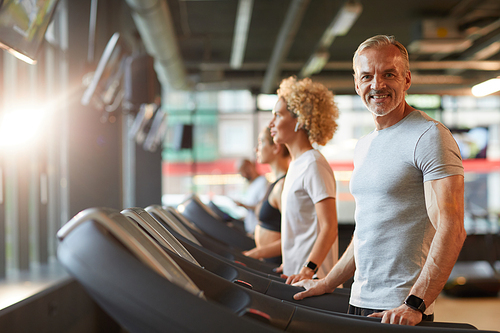 Portrait of mature man smiling at camera during his training on treadmill with other people training in the background