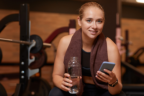 Portrait of healthy young woman with bottle of water typing a message on her mobile phone and looking at camera she resting after training