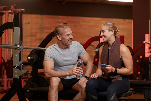 Happy couple drinking water and using mobile phone and talking to each other after sports training in gym