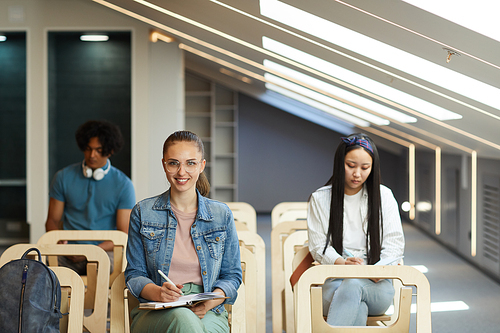 Portrait of positive student girl in denim jacket sitting on chair at lecture and making notes in diary