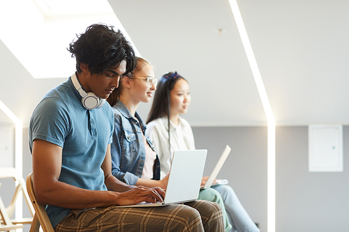 Side view of concentrated multi-ethnic students sitting in row and using laptops for notes at lecture