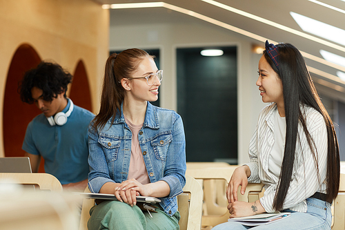 Positive multi-ethnic girls in casual outfits sitting on chairs in auditorium and chatting during break at university