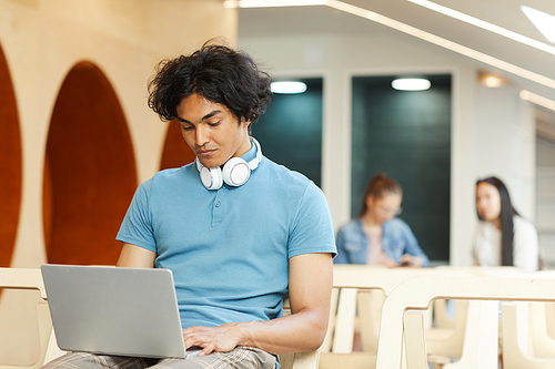 Concentrated dark-skinned student guy sitting on chair in library and using laptop while working on university project