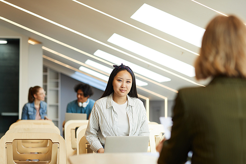 Content Asian student girl in shirt sitting in front of teacher and using laptop while passing entrance exam