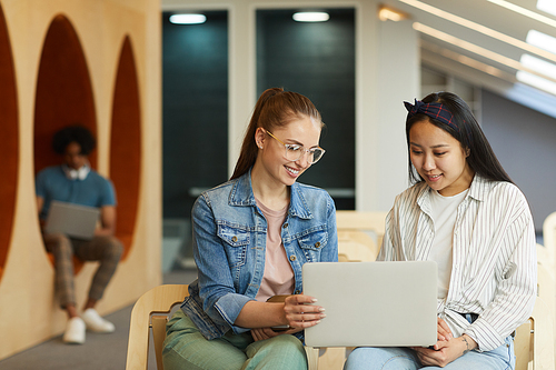 Content multi-ethnic students sitting in modern hall and working on university project together using laptop