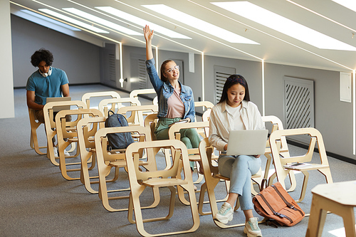 Curious student girl sitting on wooden chair and raising arm while having question at university class