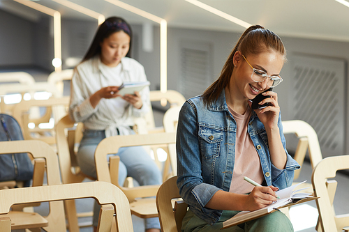 Positive attractive student girl in casual jacket sitting in university auditorium and making notes while talking by phone