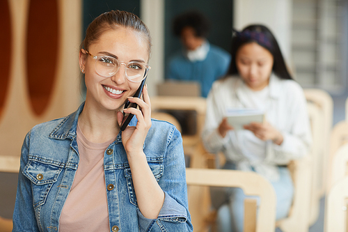 Portrait of smiling attractive student girl in eyeglasses using mobile phone in university lecture room